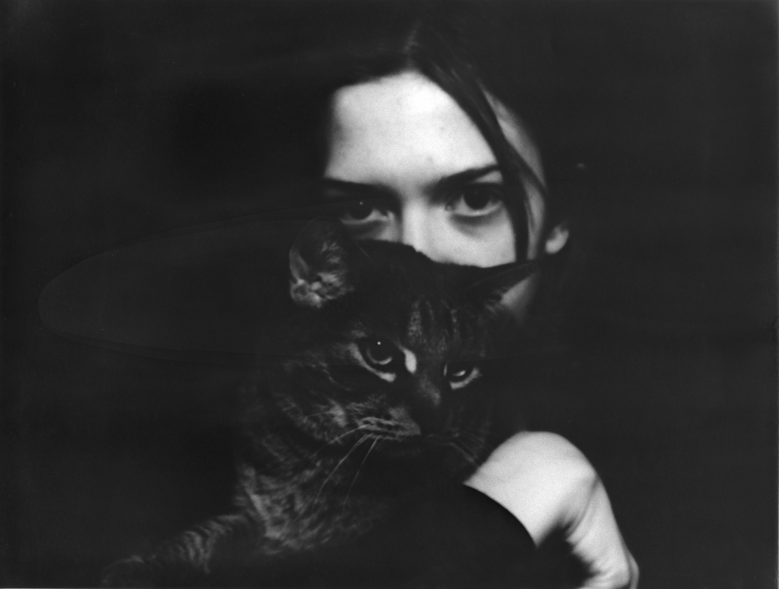 Black and white gelatin silver print of a woman whose face is partially covered by a cat that she is holding; onlye her eyes are visible. Her eyes stare accusingly out at the viewer, while the cat is distracted by something off-camera